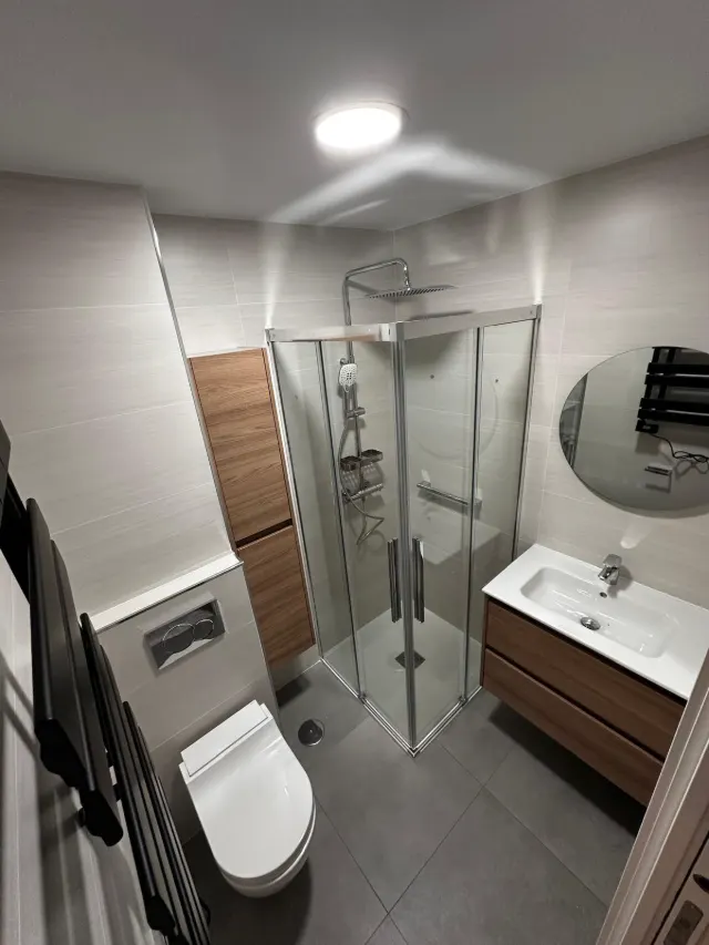 Baño reformado con plato de ducha, mampara de cristal, mueble de madera suspendido y espejo redondo